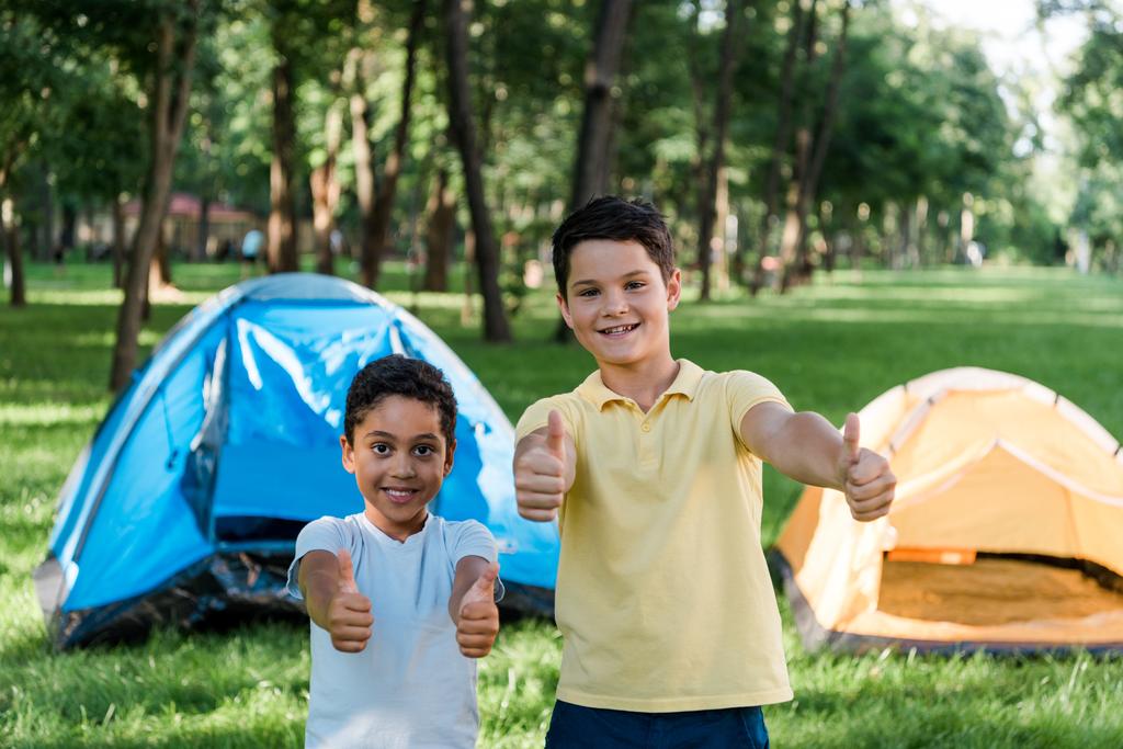 boldog multikulturális fiúk mosolyogva mutogatják hüvelykujjaikat a táborok közelében  - Fotó, kép