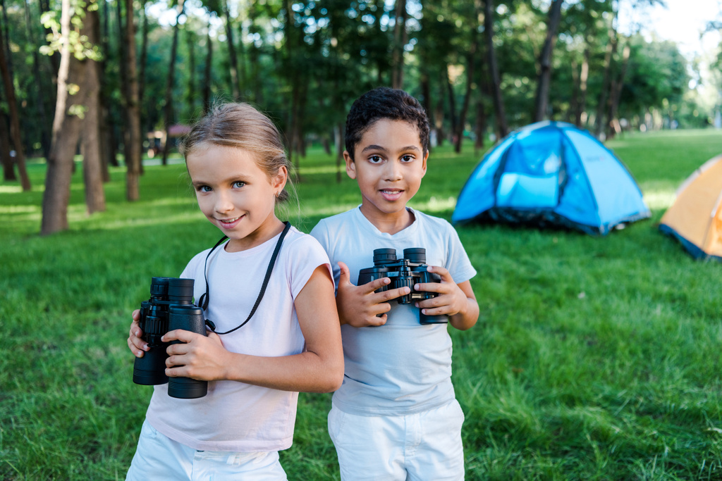 happy multicultural kids holding binoculars near camp  - Photo, Image