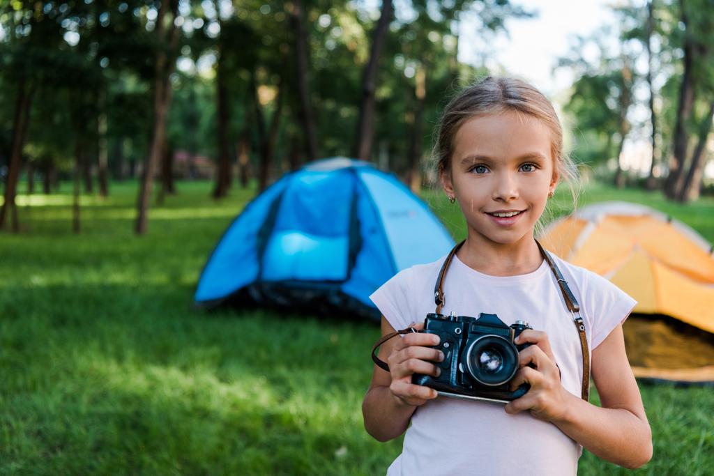 χαμογελαστό παιδί κρατώντας ψηφιακή φωτογραφική μηχανή κοντά σε στρατόπεδα στο πάρκο  - Φωτογραφία, εικόνα