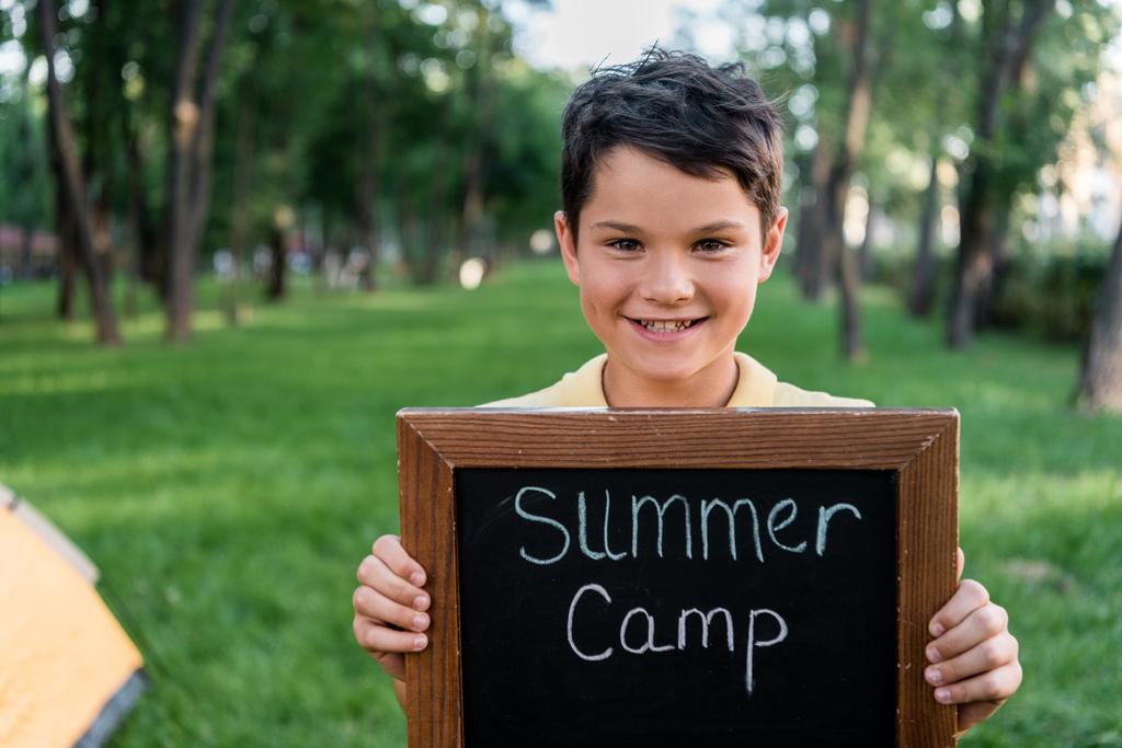 niño feliz sosteniendo pizarra con letras de campamento de verano
  - Foto, imagen
