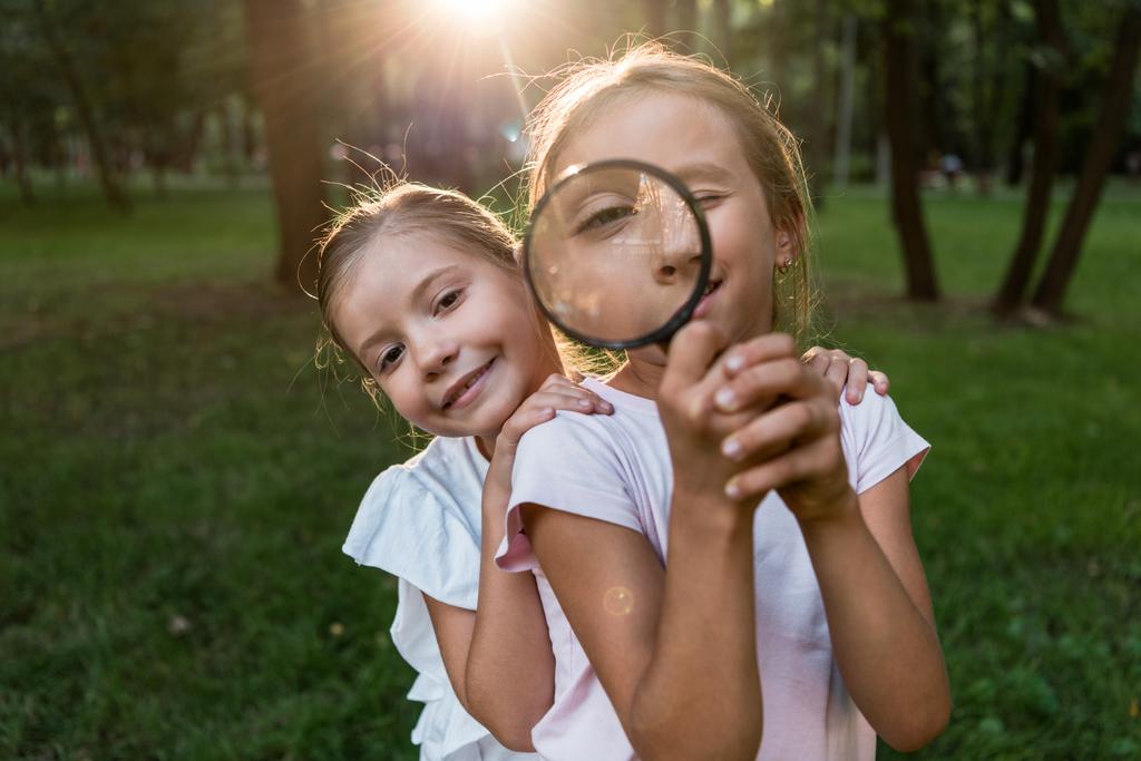 cheerful kid holding magnifier near face while standing with friend in park  - Photo, Image