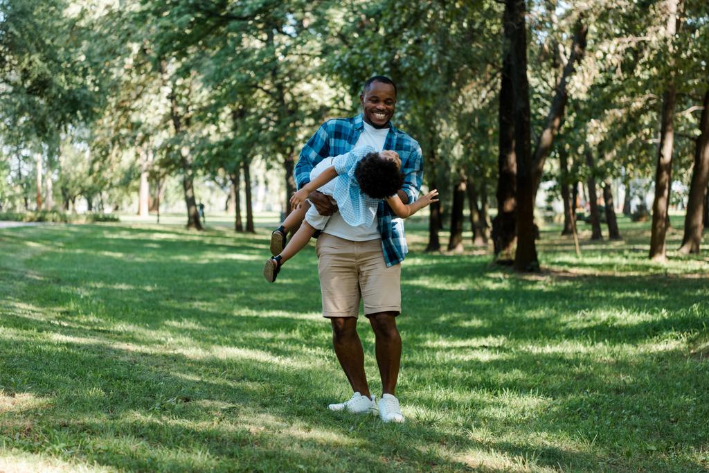 公園でかわいい息子を腕に抱きしめる陽気なアフリカ系アメリカ人の父親  - 写真・画像
