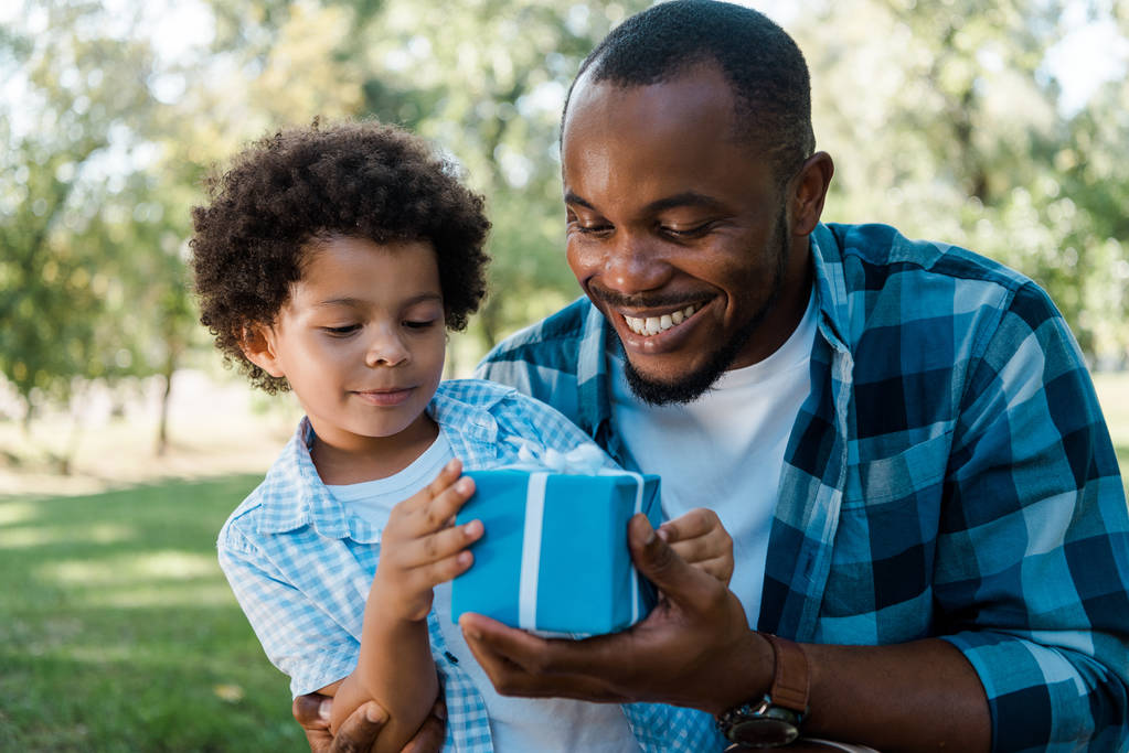 Весёлый африканский отец и сын смотрят на синюю подарочную коробку
  - Фото, изображение