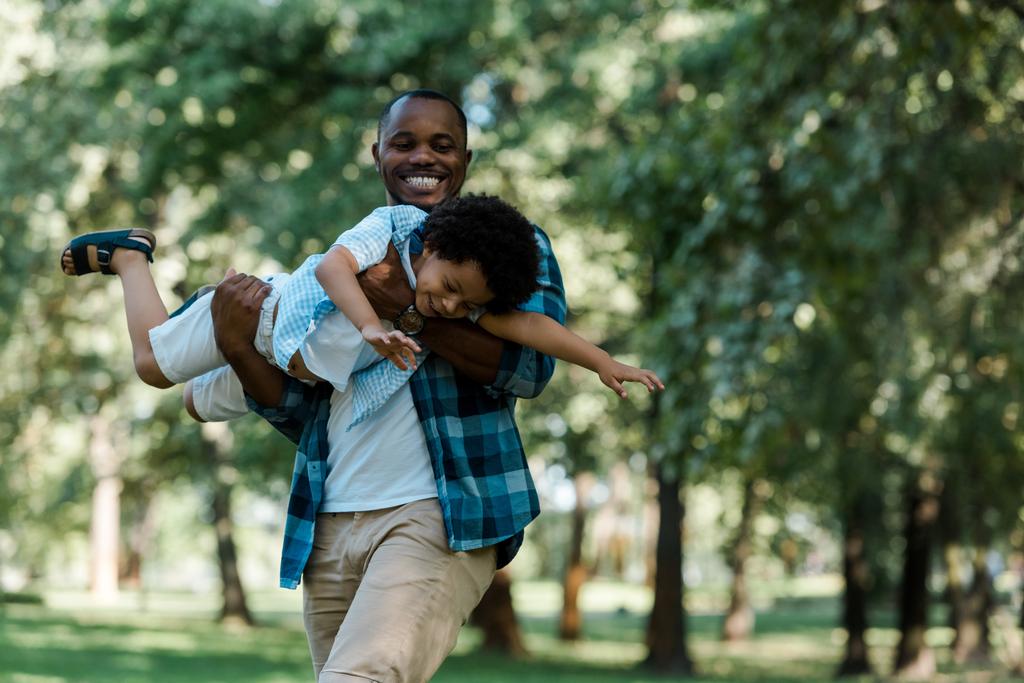 χαμογελαστός Αφρικανός Αμερικανός πατέρας κρατώντας n χέρια χαριτωμένο γιός σε πράσινο πάρκο  - Φωτογραφία, εικόνα