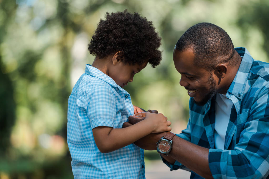 Seitenansicht des fröhlichen afrikanisch-amerikanischen Vaters beim Händchenhalten mit traurigem Sohn  - Foto, Bild