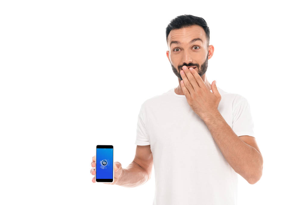 KYIV, UKRAINE - SETEMBRO 3, 2019: homem surpreso cobrindo boca e segurando smartphone com aplicativo shazam na tela isolado no branco
  - Foto, Imagem