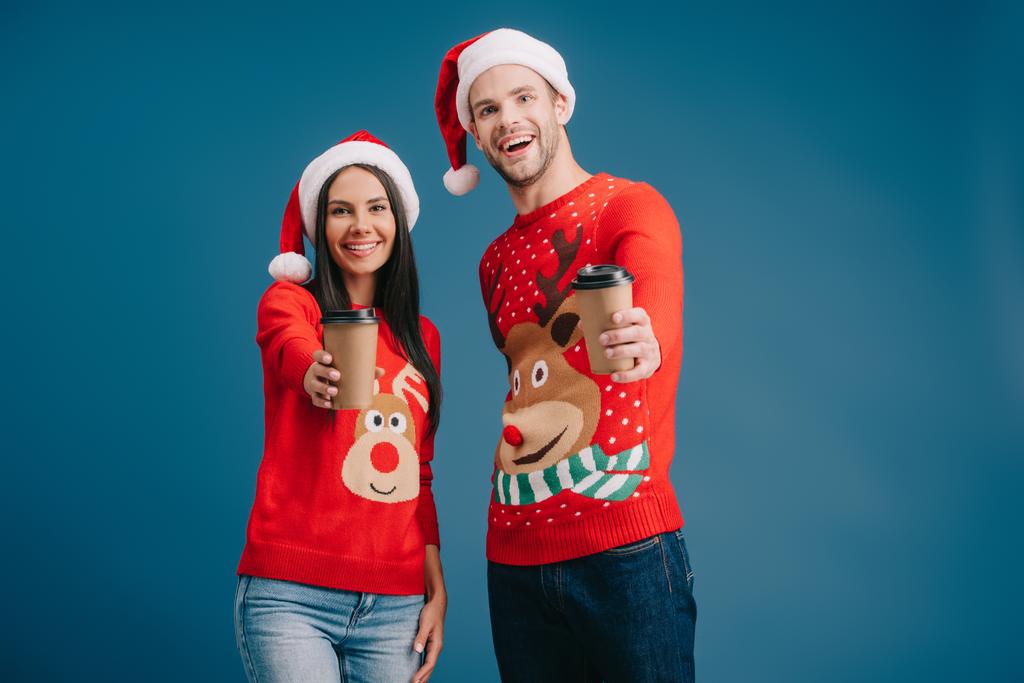 wesoły para w Santa kapelusze i Christmas Swetry trzymając kawę, aby przejść izolowane na niebiesko - Zdjęcie, obraz