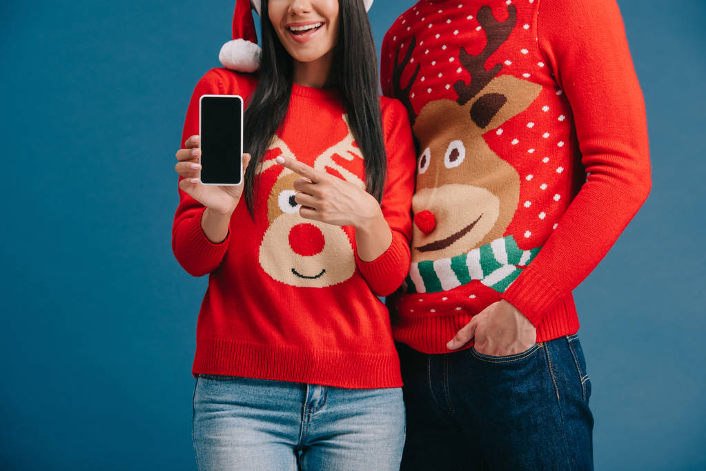 abgeschnittene Ansicht eines Paares in Weihnachtsmützen und Weihnachtspullovern, das Smartphone mit leerem Bildschirm zeigt, isoliert auf blauem Grund - Foto, Bild