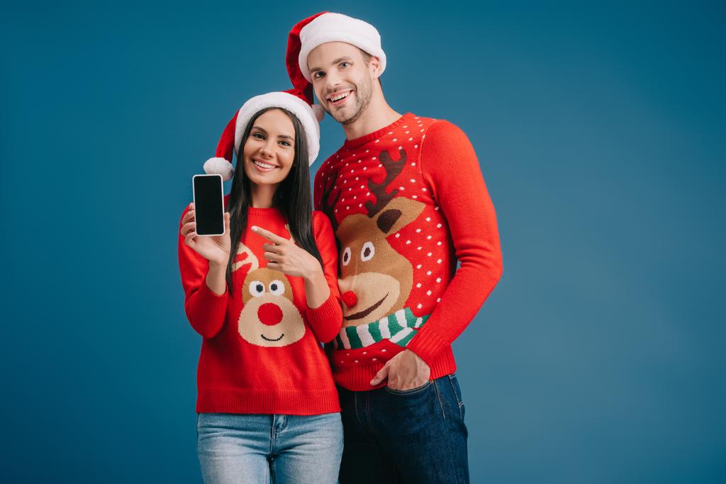 couple en chapeaux de Père Noël et pulls de Noël pointant vers le smartphone avec écran blanc isolé sur bleu
 - Photo, image