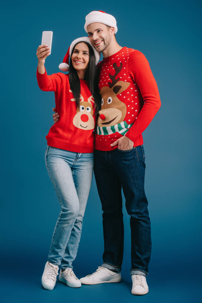couple souriant en chapeaux de Père Noël et pulls de Noël prendre selfie sur smartphone isolé sur bleu
 - Photo, image