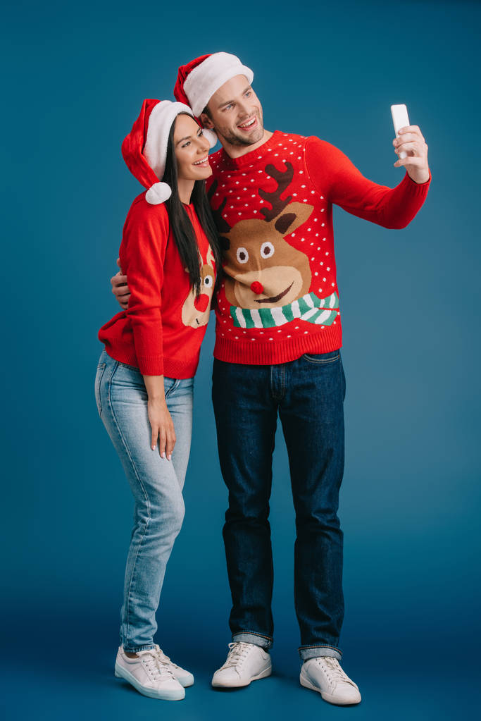 happy couple in santa hats and christmas sweaters taking selfie on smartphone isolated on blue - Photo, Image