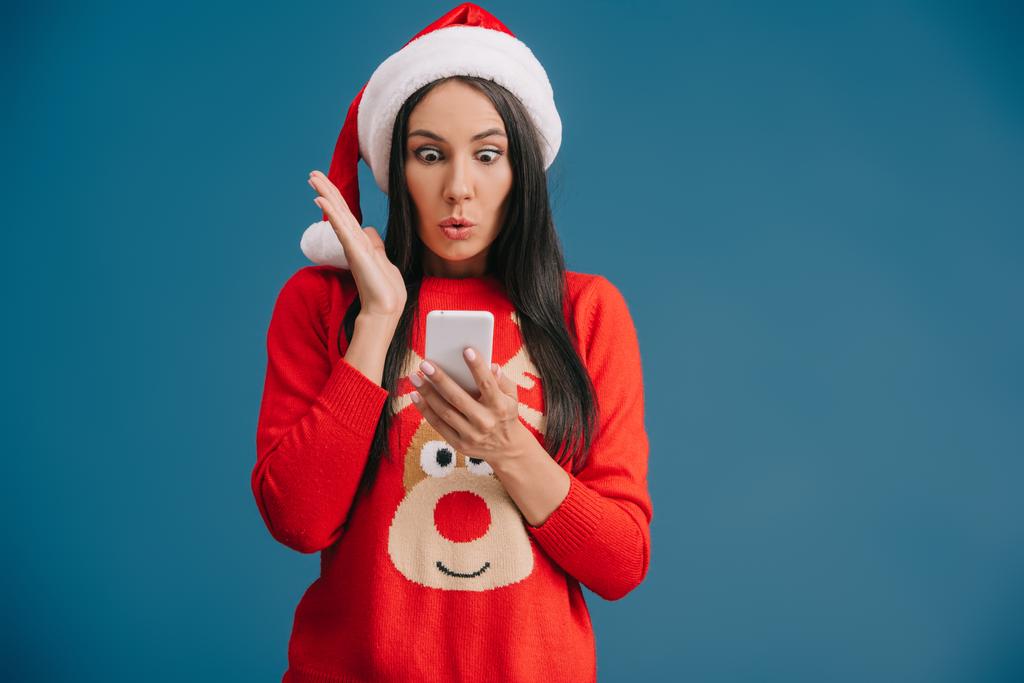 приваблива здивована жінка в капелюсі Санти та різдвяному светрі за допомогою смартфона ізольована на синьому
 - Фото, зображення