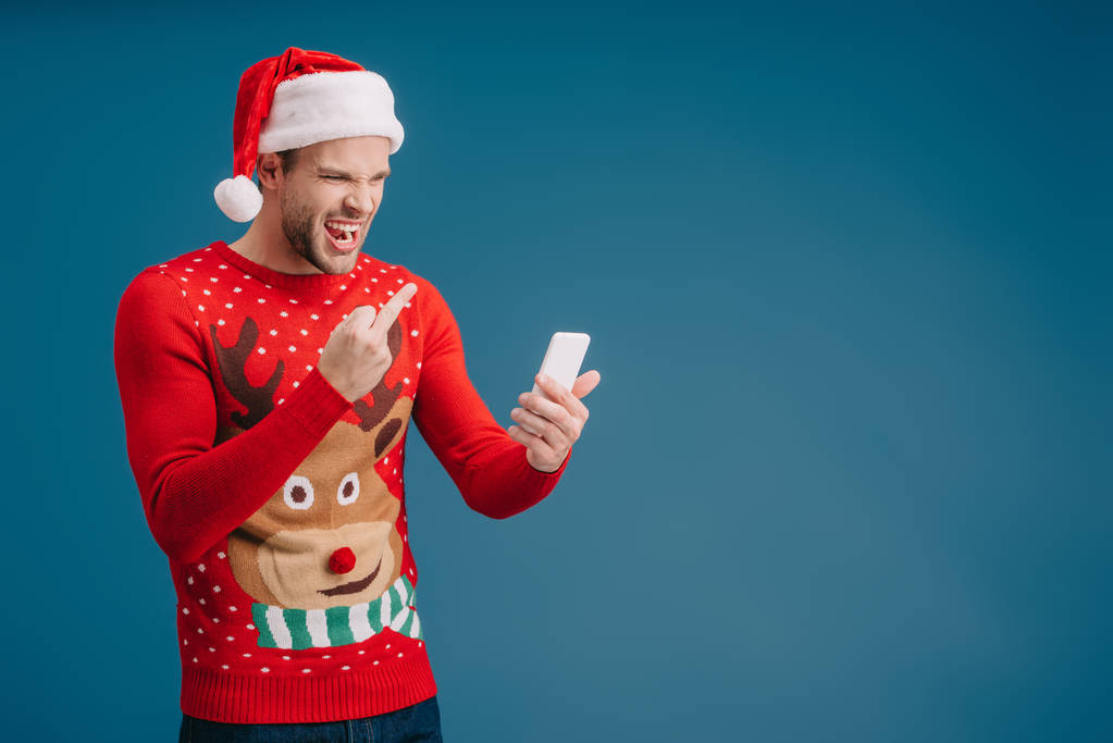Rozzlobený muž v Santa klobouku řval a ukazoval prostředníček na smartphone, izolovaný na modrém - Fotografie, Obrázek