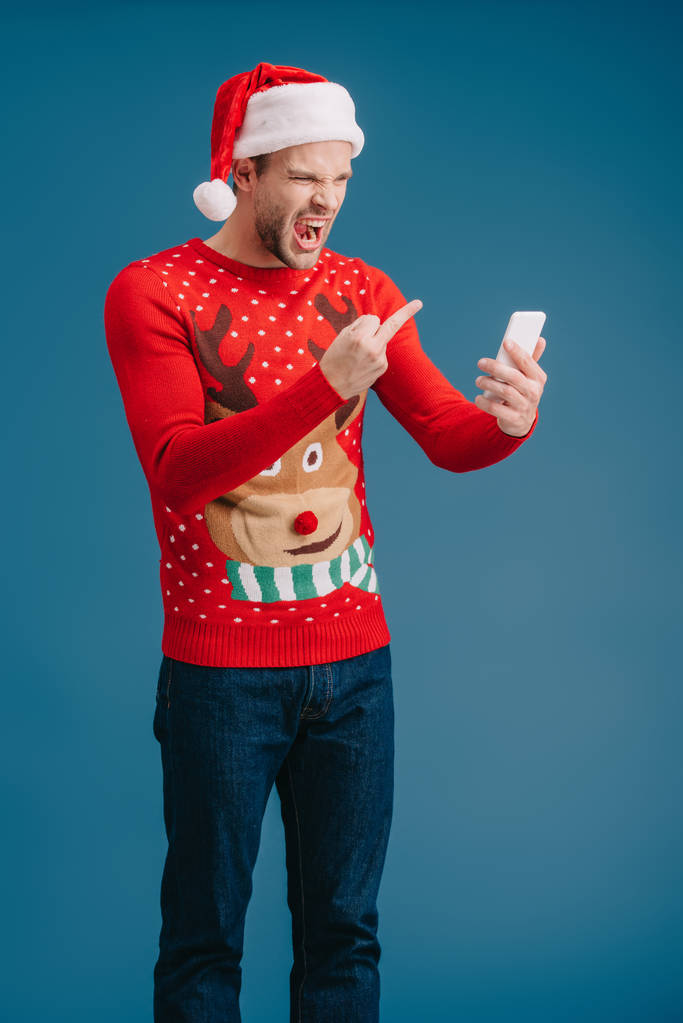 сердитий чоловік у капелюсі Санта, що кричить і показує середній палець на смартфон, ізольований на синьому
 - Фото, зображення