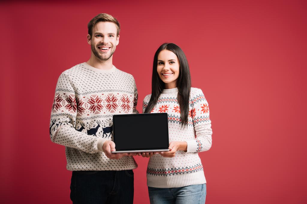 όμορφο ζευγάρι σε χειμωνιάτικα πουλόβερ παρουσιάζοντας φορητό υπολογιστή με κενή οθόνη απομονωθεί στο κόκκινο - Φωτογραφία, εικόνα