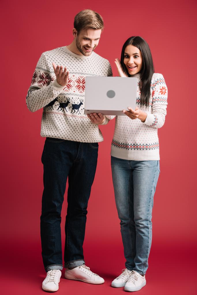 překvapené dvojice v zimních svetry používající přenosný počítač izolovaný na červenou - Fotografie, Obrázek