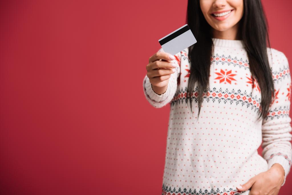 クレジットカードを保持する冬のセーターの女性のトリミングビュー, 赤で隔離  - 写真・画像