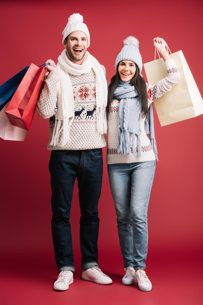 cheerful couple in winter sweaters, scarves and hats holding shopping bags isolated on red - Photo, Image