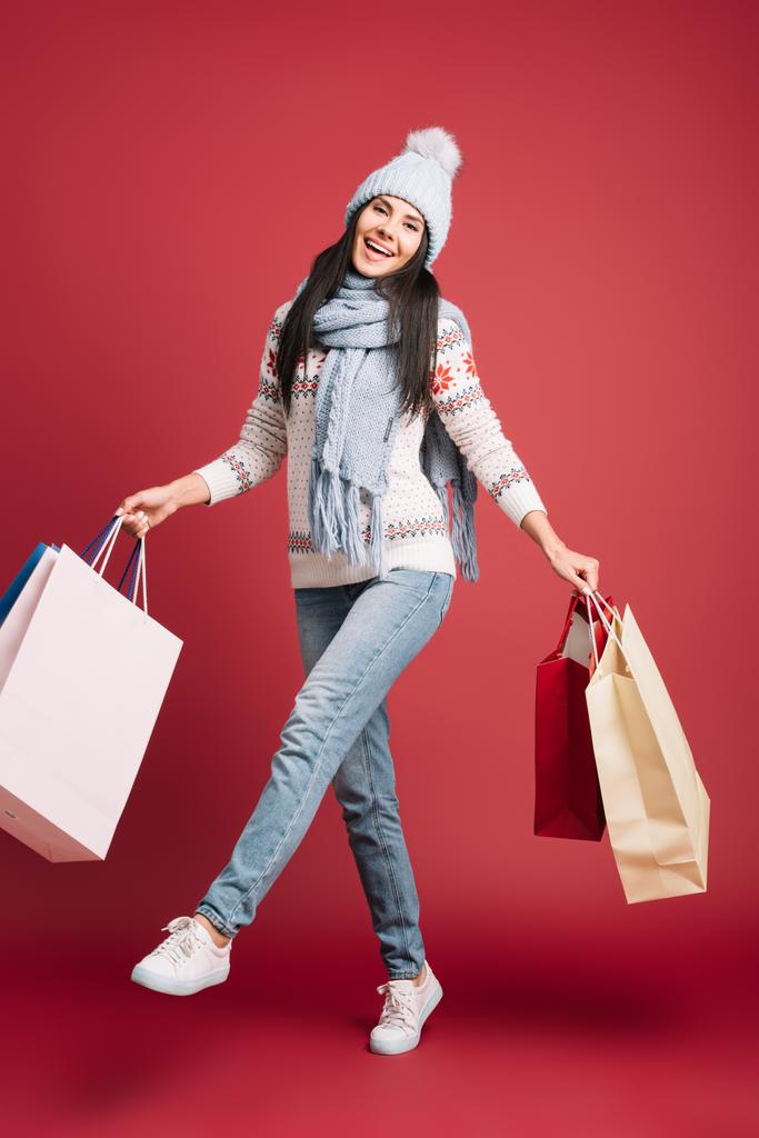 bella ragazza in maglione invernale, sciarpa e cappello in possesso di borse della spesa, isolato su rosso
  - Foto, immagini