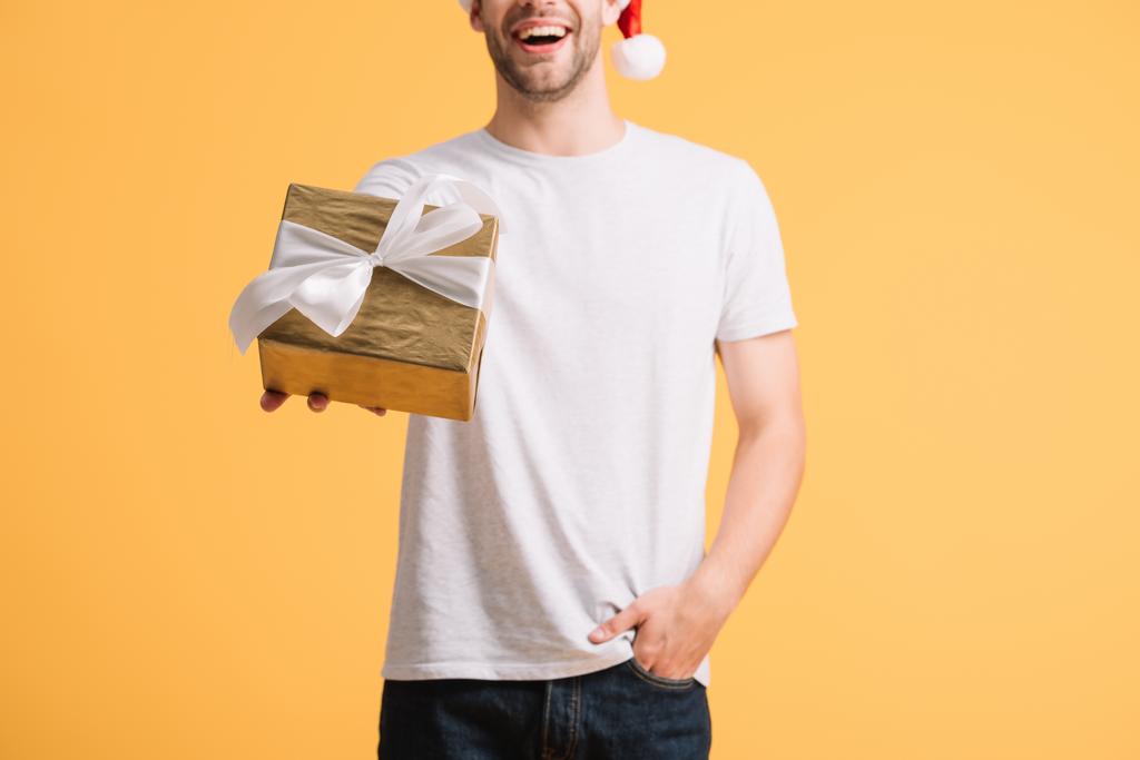 黄色に隔離されたクリスマスプレゼントを保持するサンタ帽子の幸せな男のトリミングされたビュー - 写真・画像