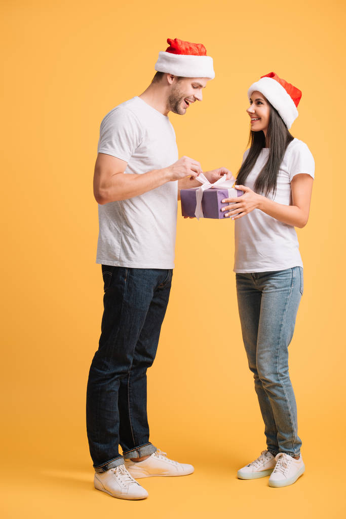 ζευγάρι σε Σάντα καπέλα άνοιγμα χριστουγεννιάτικο δώρο απομονωμένο στο κίτρινο - Φωτογραφία, εικόνα