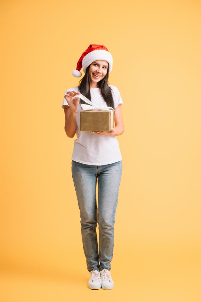 красива усміхнена дівчина в капелюсі Санта тримає подарунок Різдва ізольовано на жовтому
 - Фото, зображення