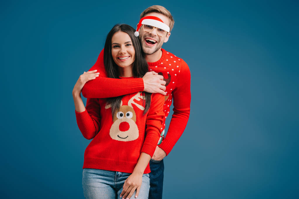 cheerful man in glasses with santa hat hugging woman isolated on blue - Photo, Image