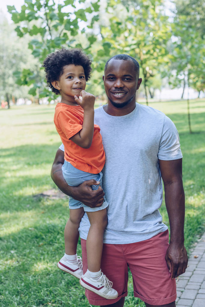 heureux homme afro-américain tenant adorable fils et regardant la caméra tout en marchant dans le parc
  - Photo, image