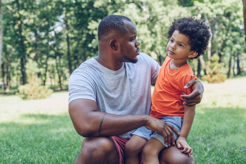 cute african american boy sitting on fathers knee in park - Photo, Image
