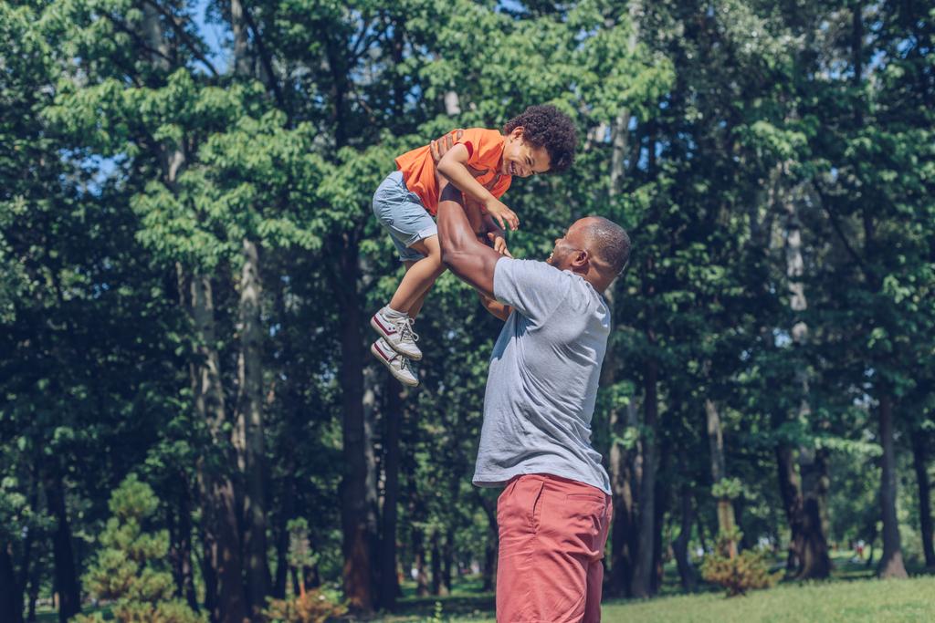 jovem afro-americano homem segurando alegre filho acima da cabeça enquanto se divertindo no parque
 - Foto, Imagem