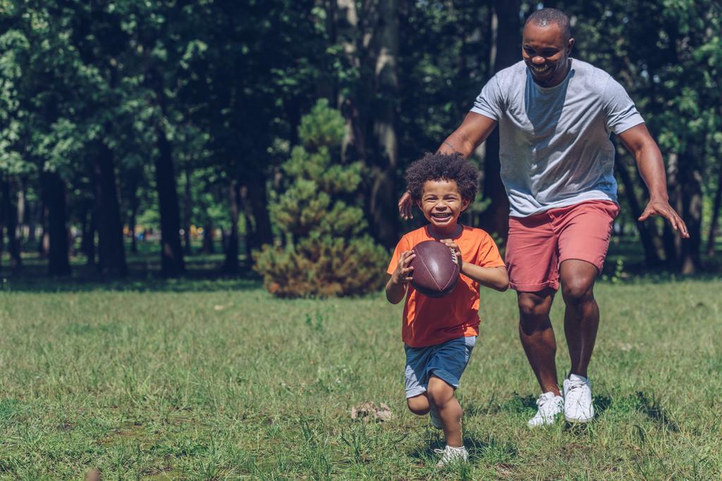 公園で父親と遊びながらラグビーボールで走る可愛いアフリカ系アメリカ人少年 - 写真・画像