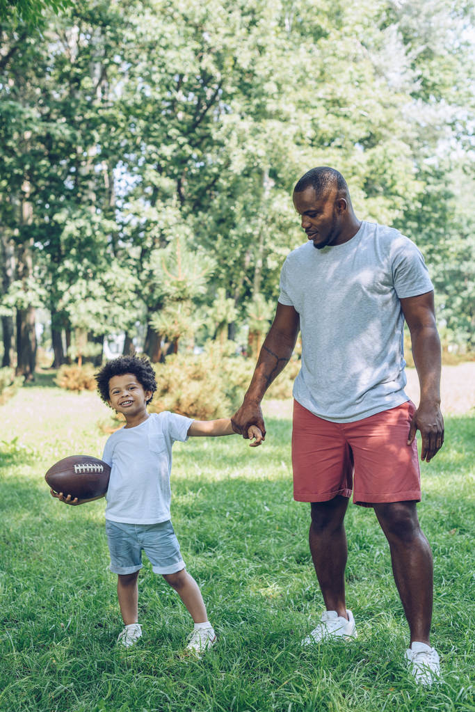 χαριτωμένο αφροαμερικανός αγόρι hoding μπάλα ράγκμπι, ενώ κρατώντας το χέρι με τον πατέρα στο πάρκο - Φωτογραφία, εικόνα