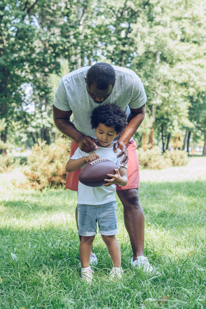 jong Afrikaans amerikaans man knuffelen zoon holding rugby bal in park - Foto, afbeelding