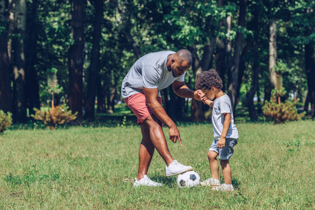 νεαρός Αφροαμερικάνος διδάσκει αξιολάτρευτο γιο παίζοντας ποδόσφαιρο στο πάρκο - Φωτογραφία, εικόνα