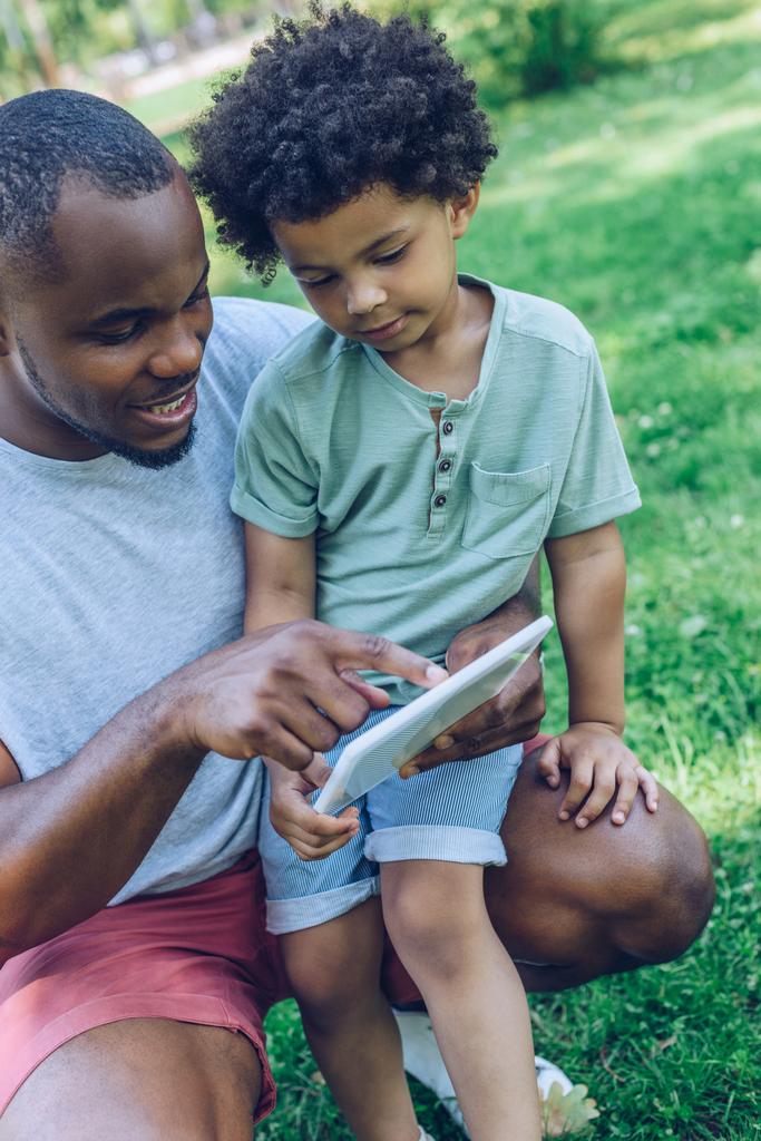 Lächelnder Afroamerikaner zeigt seinem entzückenden Sohn im Park digitales Tablet - Foto, Bild