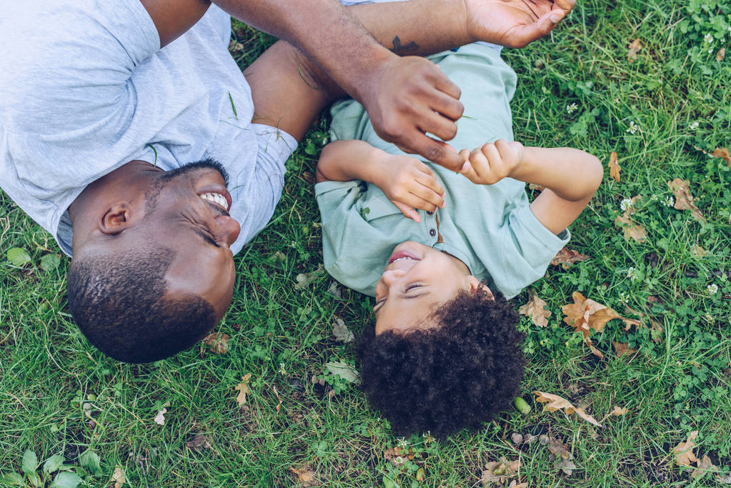 overhead view of african american father and son having fun while lying on lawn in park - Photo, Image