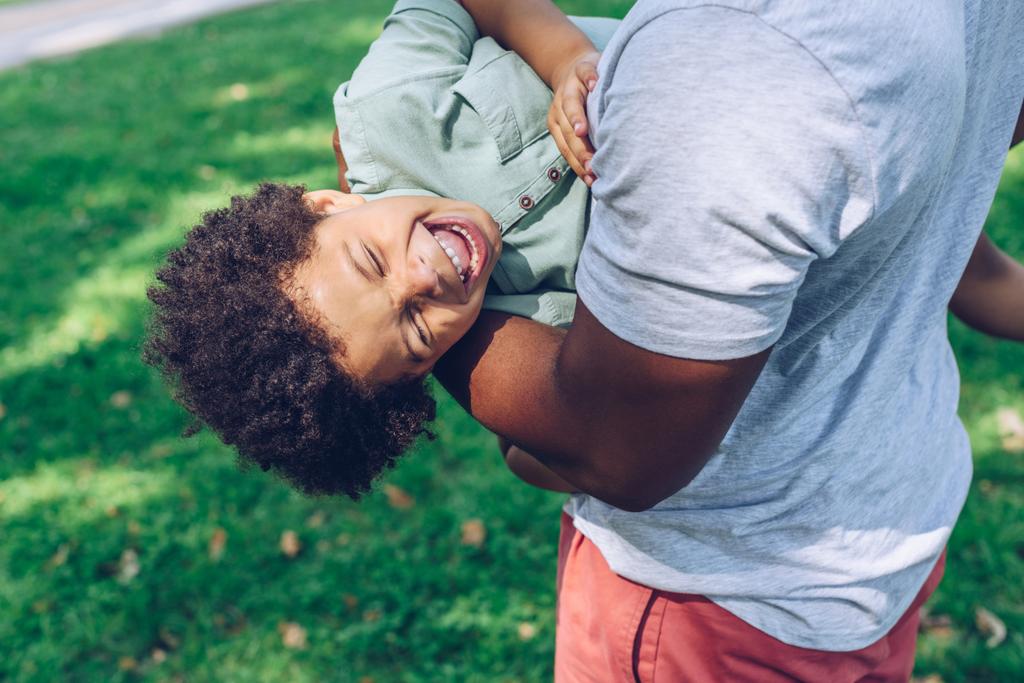 cropped view of african american man holding laughing son while having son in park - Photo, Image