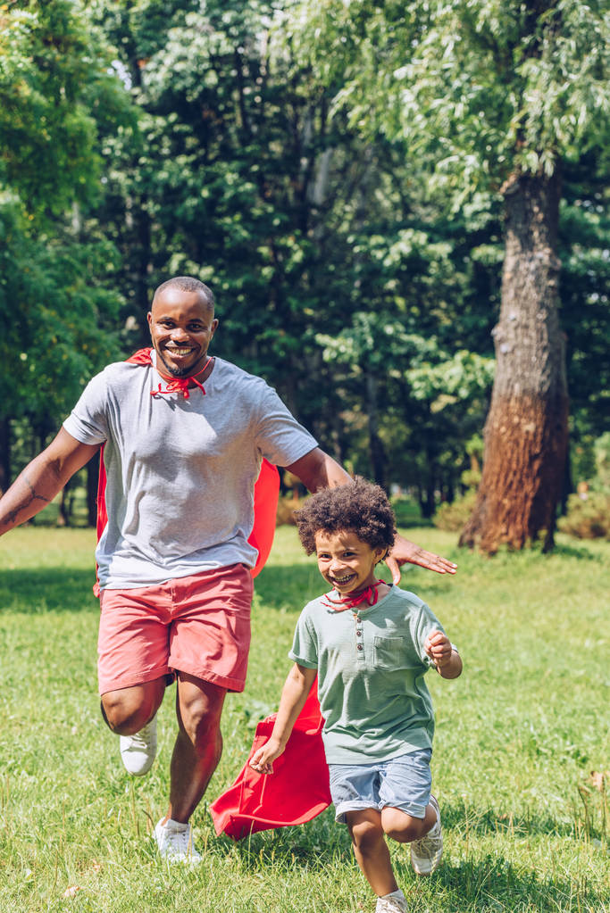 cheerful african american father and son running in costumes of superheroes in park - Photo, Image