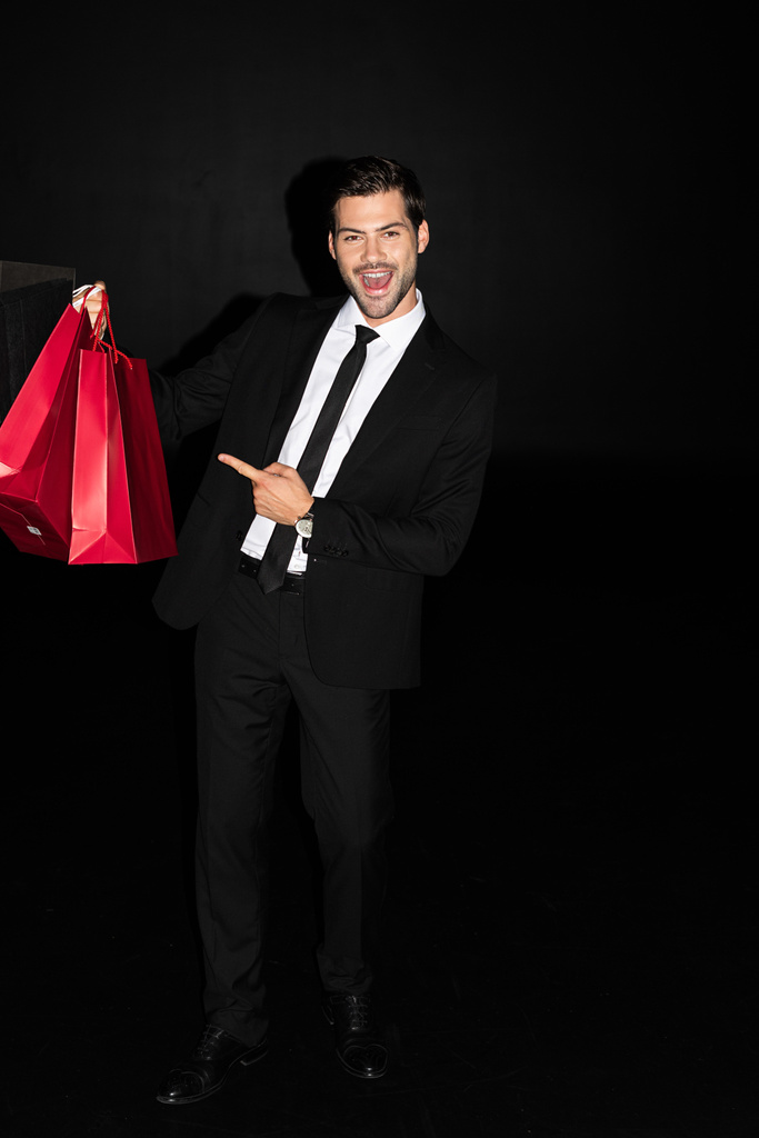 елегантний збуджений чоловік у костюмі, що показує червоні сумки для покупок ізольовані на чорному
 - Фото, зображення