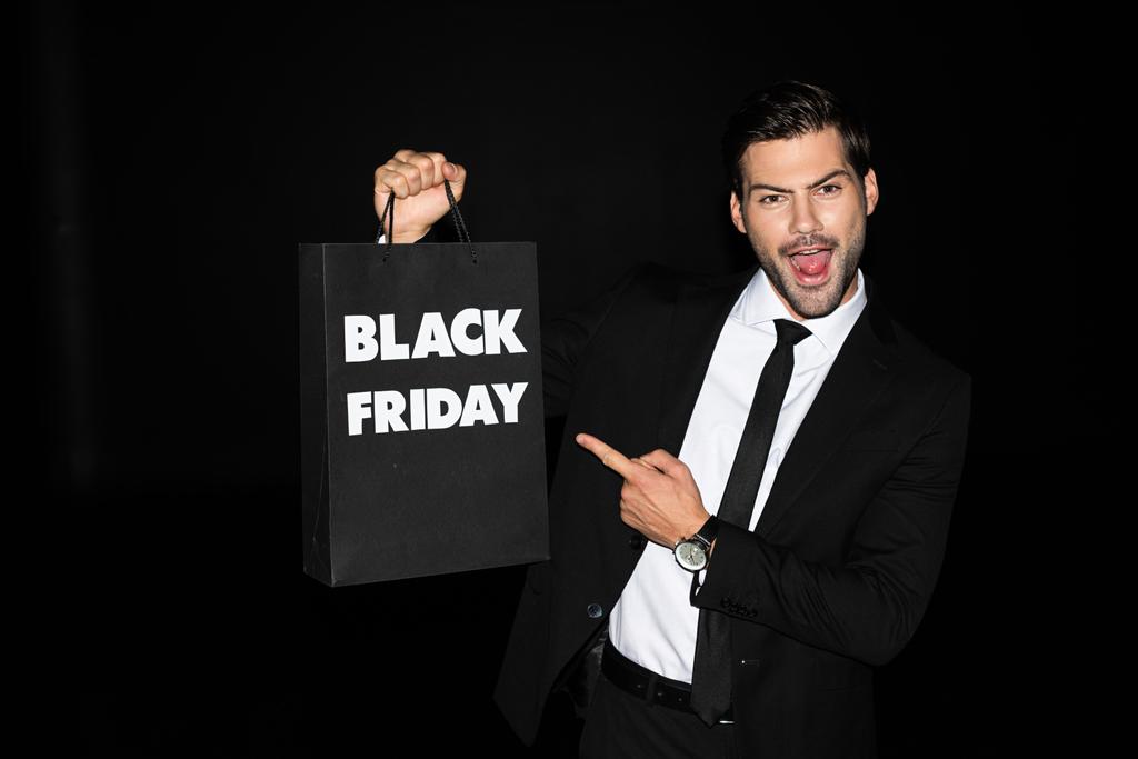 vzrušený muž ukazující nákupní tašku s černým pátečním znakem, izolovaný na černém - Fotografie, Obrázek