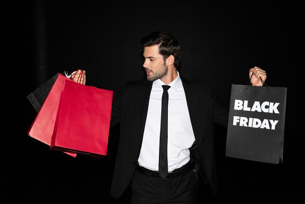 elegante hombre sosteniendo bolsas de compras con signo de viernes negro, aislado en negro
  - Foto, Imagen