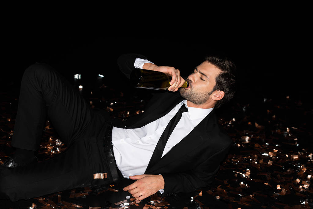 elegante uomo bere champagne dalla bottiglia mentre sdraiato sul pavimento con confetti d'oro per le vacanze
 - Foto, immagini