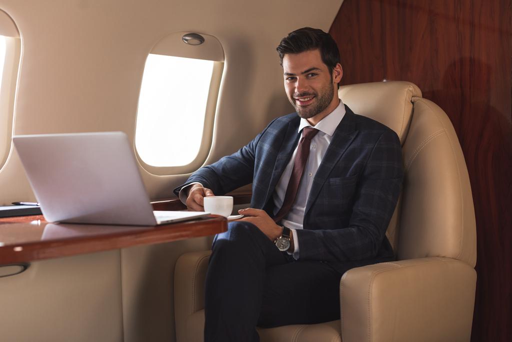 glimlachende zakenman houdt kopje koffie in vliegtuig met laptop tijdens zakenreis  - Foto, afbeelding