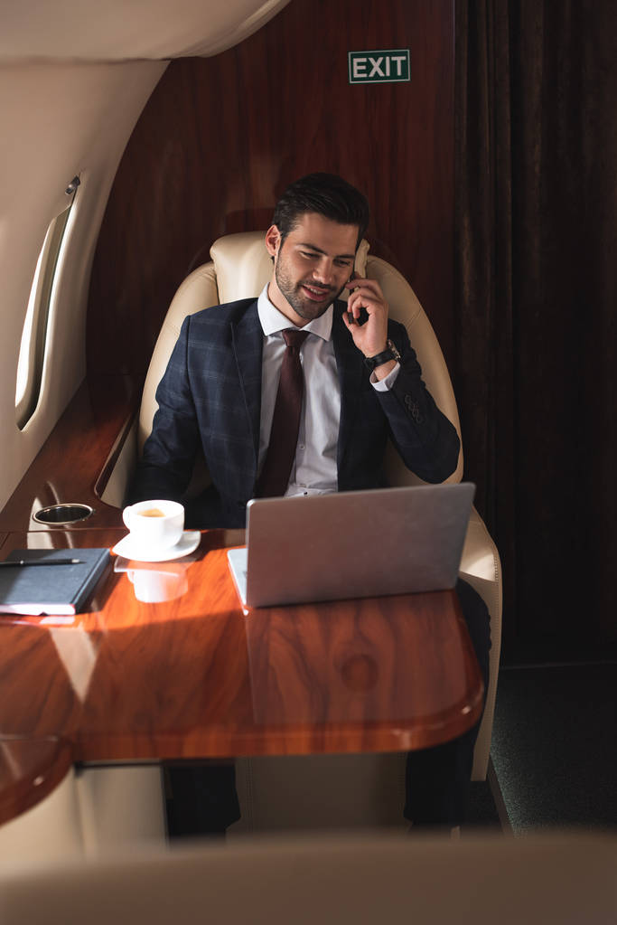 glimlachende zakenman praten op smartphone in vliegtuig met laptop tijdens zakenreis  - Foto, afbeelding