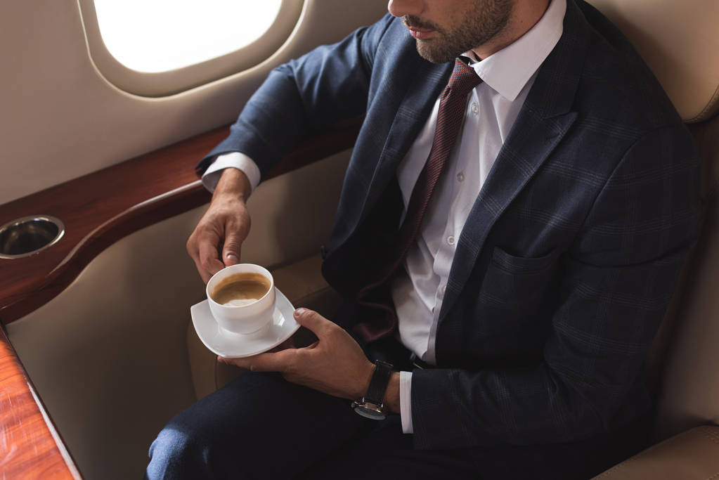 bijgesneden beeld van zakenman in pak met koffiebeker in vliegtuig tijdens zakenreis - Foto, afbeelding
