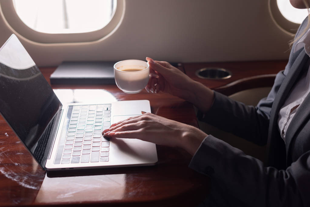 cropped view of businesswoman holding coffee cup and working on laptop in plane during business trip  - 写真・画像