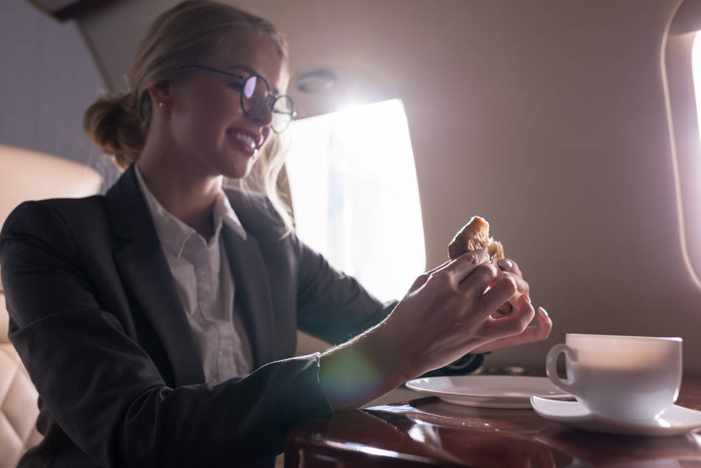 ελκυστική επιχειρηματίας πίνοντας καφέ με κρουασάν στο αεροπλάνο κατά τη διάρκεια επαγγελματικού ταξιδιού - Φωτογραφία, εικόνα