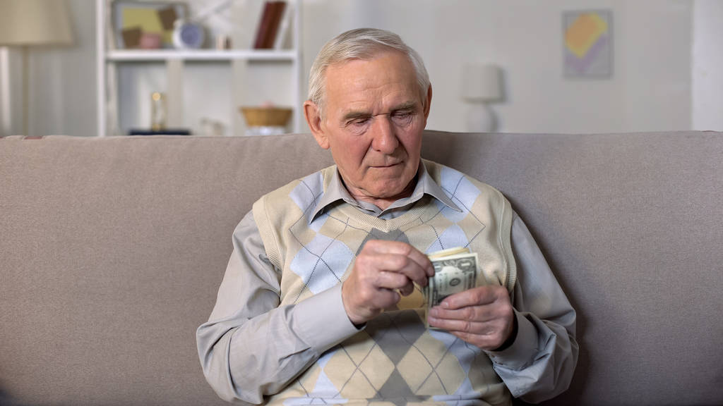 Depressed old man sitting on sofa holding dollars banknotes, social insecurity - Foto, afbeelding
