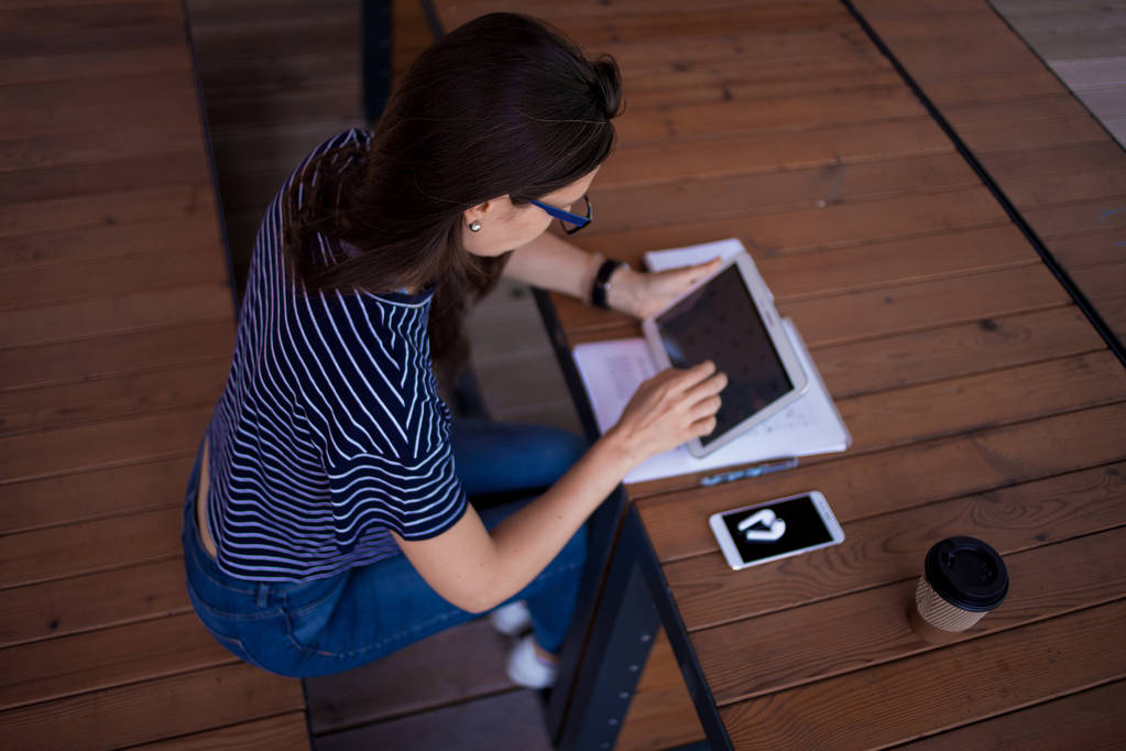 Une fille brune sérieuse, pigiste, avec une grande montre-bracelet, travaille à une table en bois sur tablette, avec des gadgets et des papiers
. - Photo, image