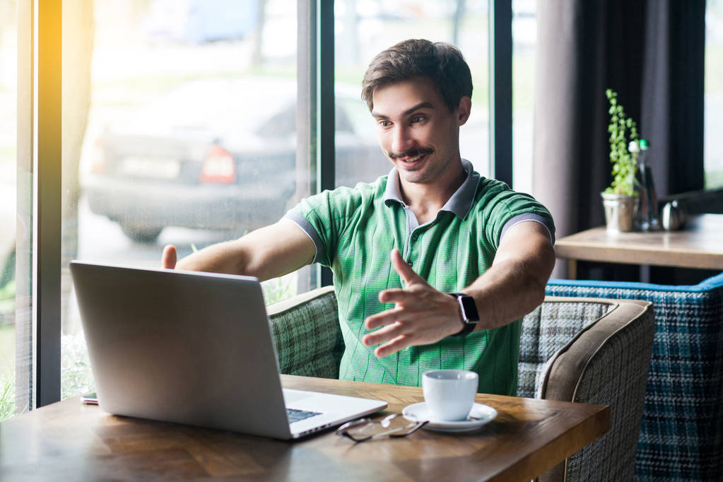 Young happy businessman in green t-shirt looking at laptop on video call and showing embrace gesture while sitting in cafe, freelancing concept - Photo, Image